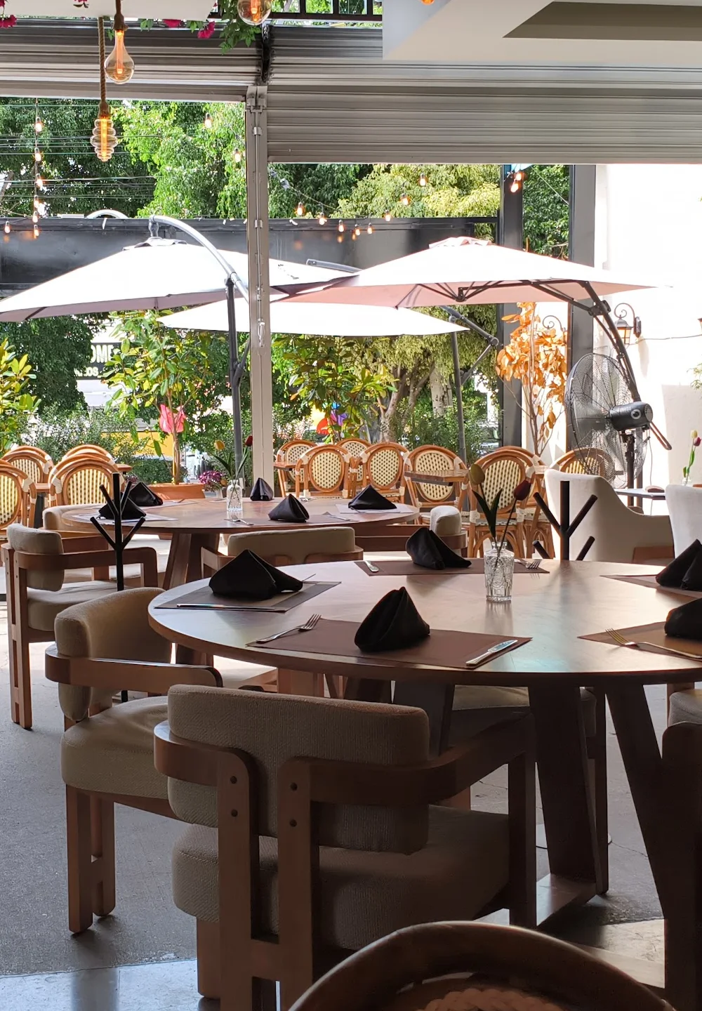 La terraza de Querena vista desde el salón: sombrillas blancas, plantas, mesas de madera con servilletas dobladas y focos de cuerda colgados.