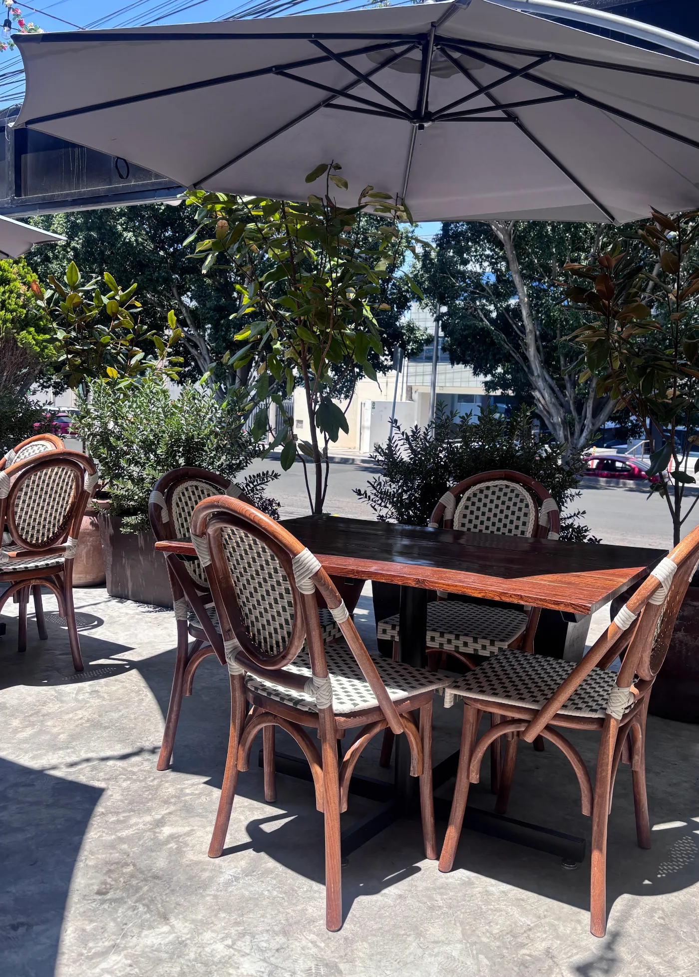 La terraza de Querena bajo una sombrilla blanca: mesa de madera, sillas de bejuco y los árboles del boulevard detrás.