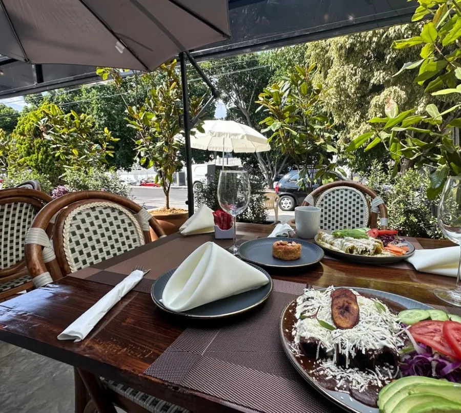 La terraza de Querena por la mañana: mesa de madera puesta con platos de desayuno, copas y jugo, bajo la sombrilla y entre las plantas.
