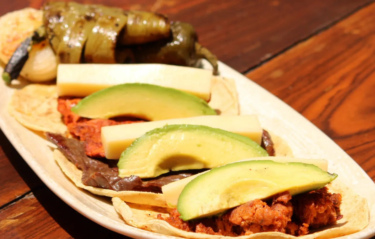 Tacos de cecina de Yecapixtla vistos de cerca: láminas de aguacate y queso sobre la carne, en un plato ovalado encima de la mesa de madera.