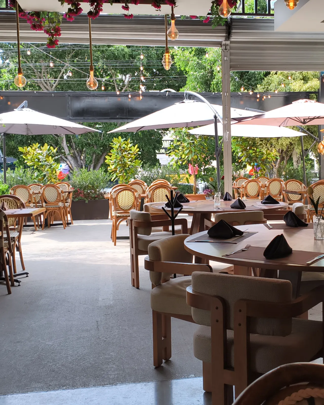 El comedor cubierto de Querena abierto a la terraza a plena luz: mesas redondas puestas con servilletas negras, sillas de bejuco, sombrillas al fondo y la bugambilia colgando del techo.