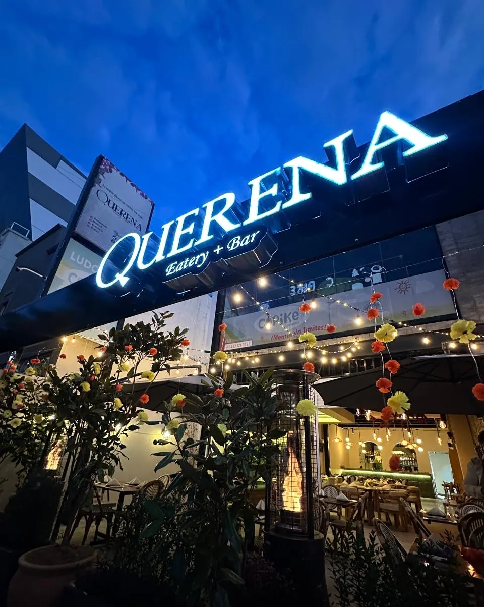 La fachada de Querena al caer la tarde, con el letrero iluminado contra el cielo azul y las luces de la terraza encendidas.