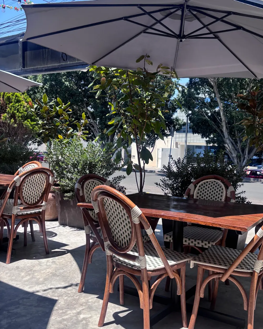 La terraza de Querena de día: mesa de madera bajo una sombrilla blanca, sillas de bejuco y árboles alrededor.