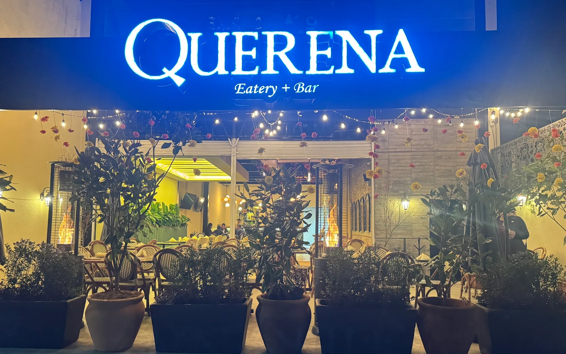 El letrero iluminado de Querena contra el cielo del anochecer, con la hilera de macetas, los focos de cuerda y el interior encendido detrás.