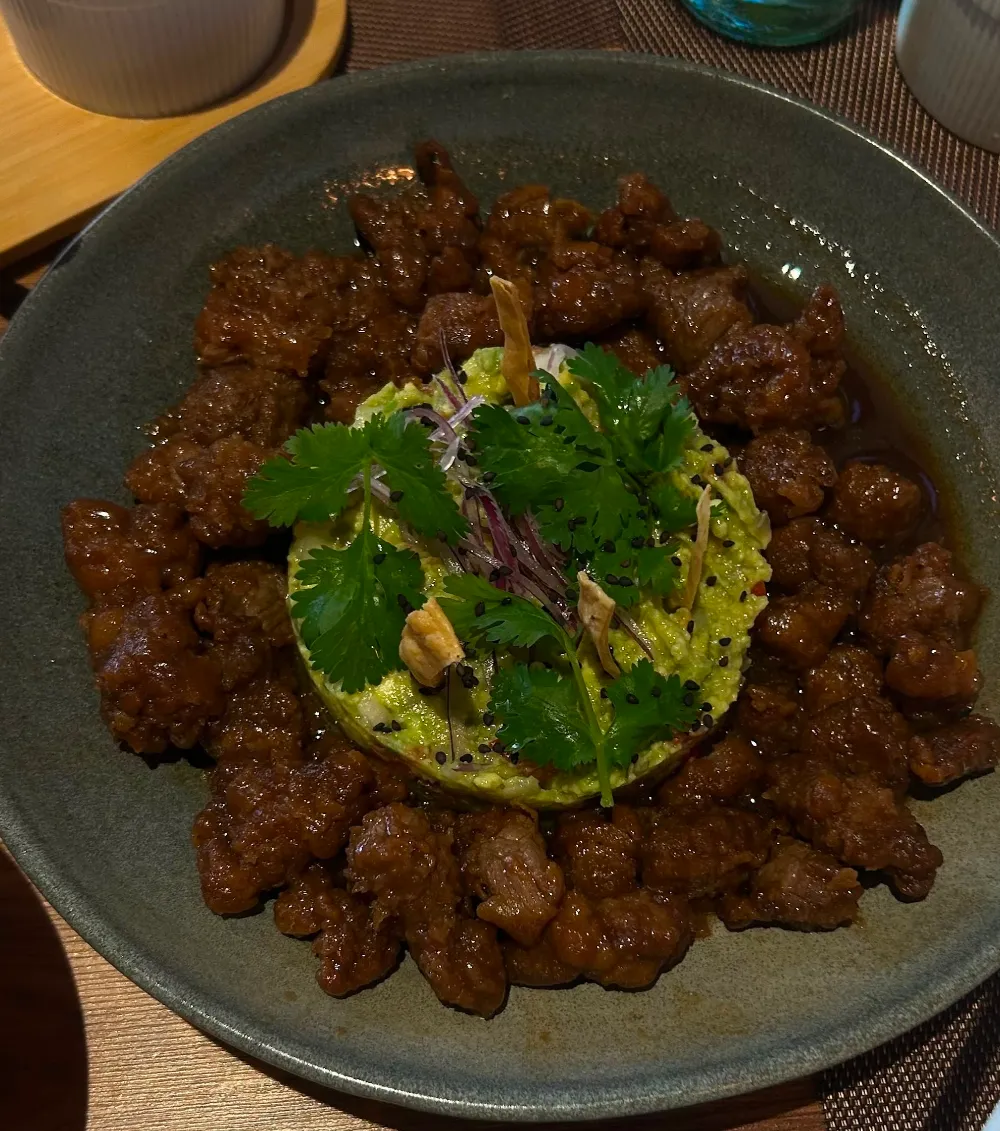Bowl oscuro con trozos de carne, guacamole al centro y hojas verdes alrededor.