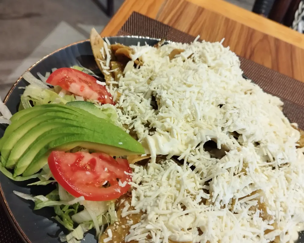 Chilaquiles verdes cubiertos de queso rallado, con láminas de aguacate, jitomate y col, en un plato oscuro.