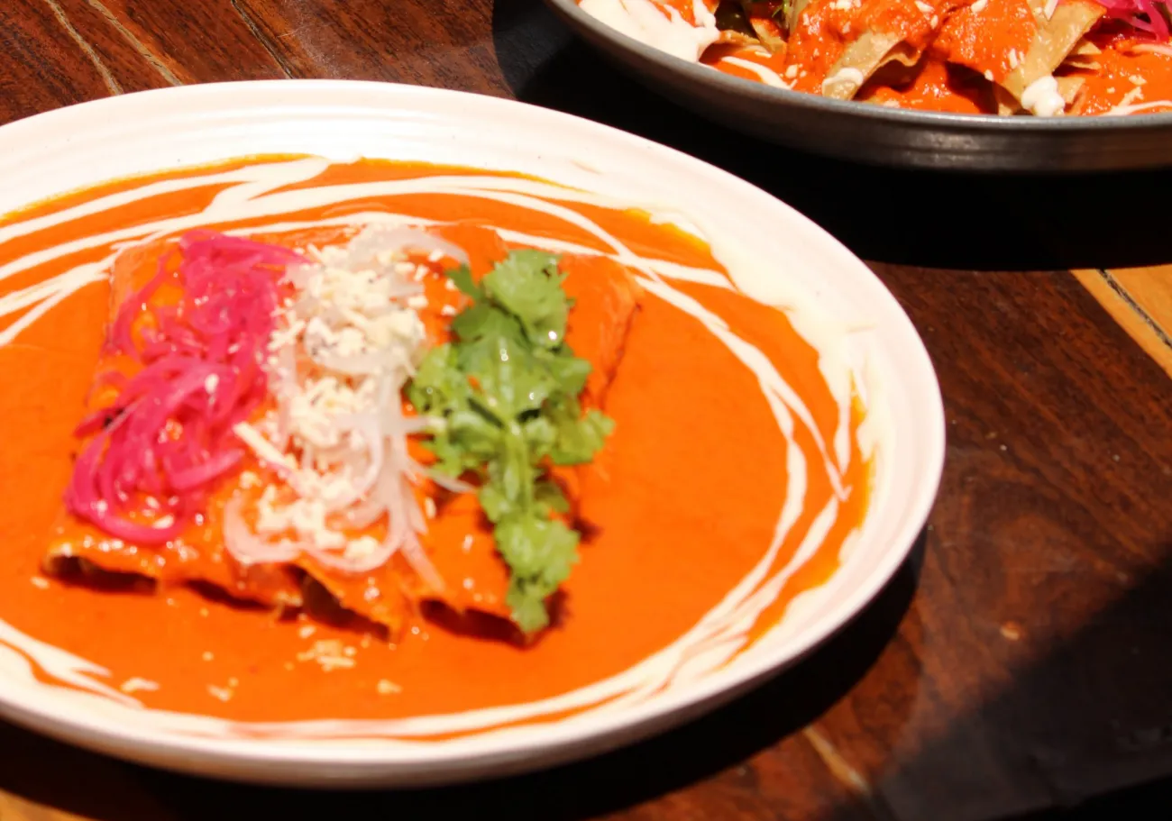 Dos platos servidos sobre una mesa de madera: enchiladas en salsa roja al frente y un tazón de chilaquiles detrás.