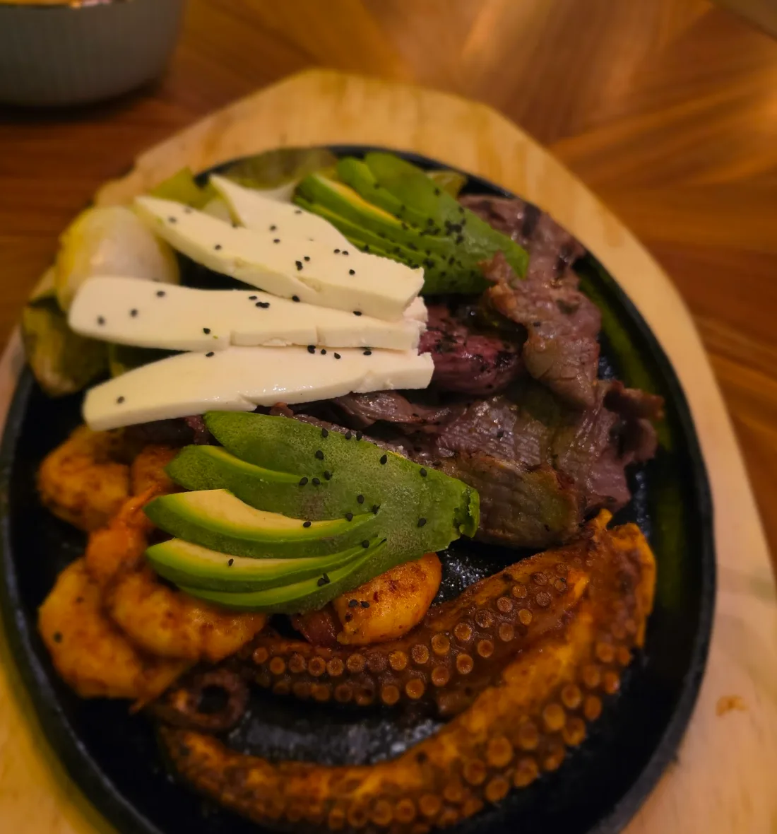 Comal negro con carne, tentáculos de pulpo, tiras de queso blanco, aguacate y plátano frito, sobre una tabla de madera.