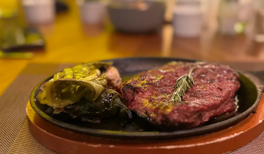 Un corte de carne sobre un comal de barro con chiles asados, y el salón desenfocado al fondo.