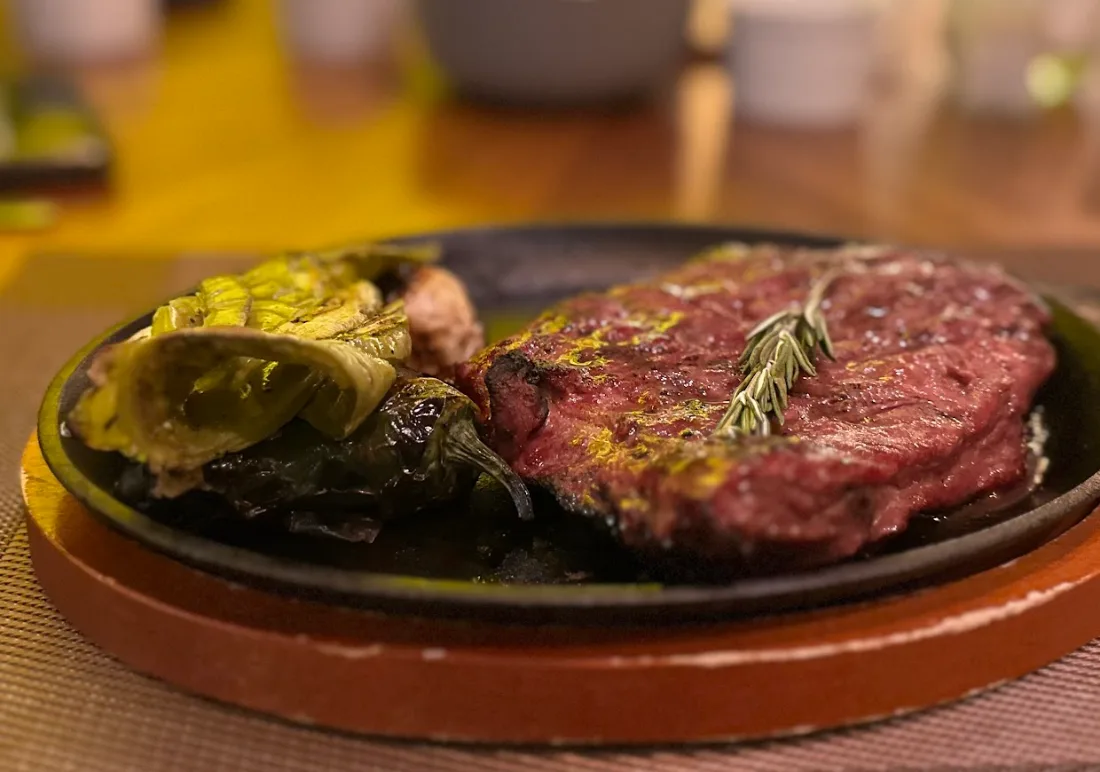 Un corte de carne sobre un comal de barro con chiles asados, y el salón desenfocado al fondo.
