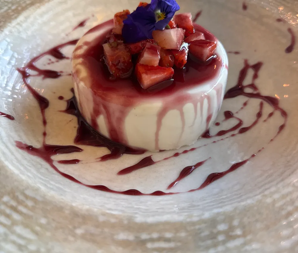 Postre blanco montado con fruta picada y una flor comestible morada, sobre reducción de frutos rojos.