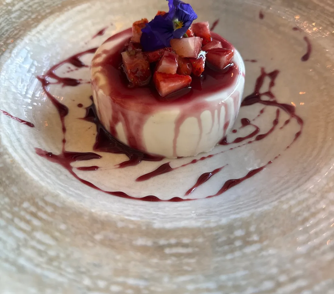 Postre blanco montado con fruta picada y una flor comestible morada, sobre un plato de cerámica con reducción de frutos rojos.