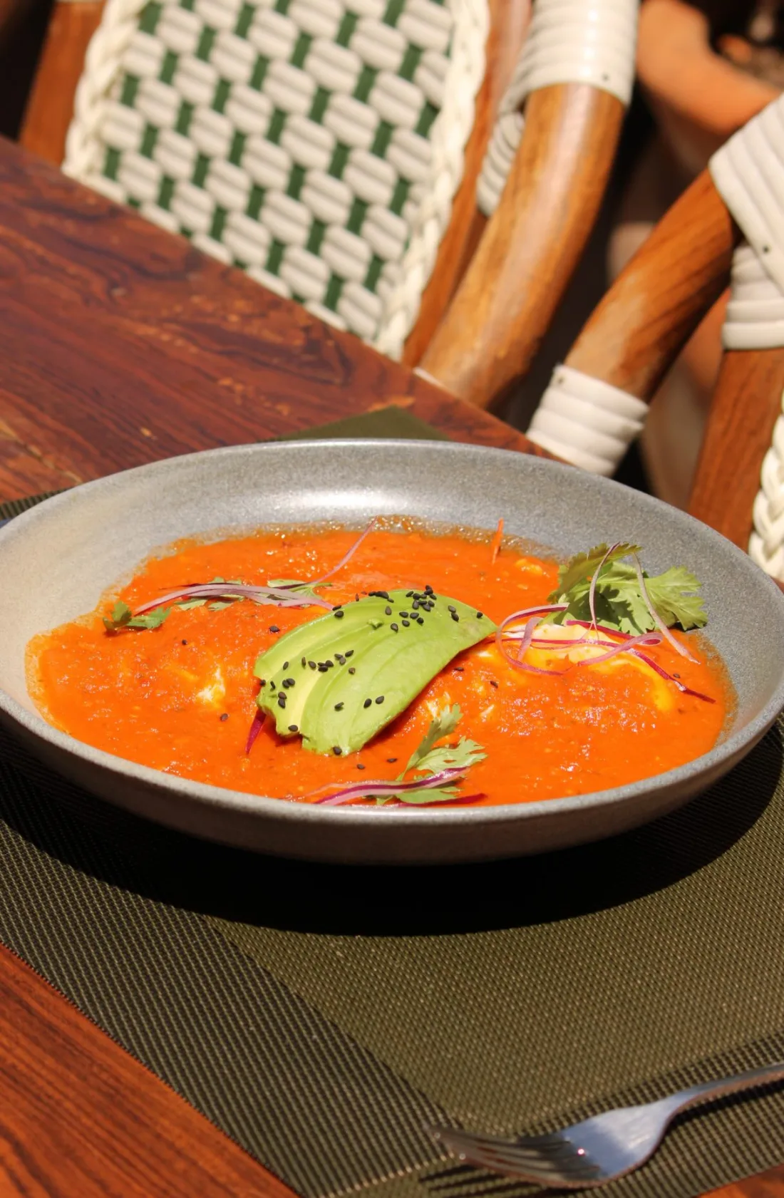 Huevo sobre salsa roja con láminas de aguacate y ajonjolí negro, en un plato hondo sobre una mesa de madera.