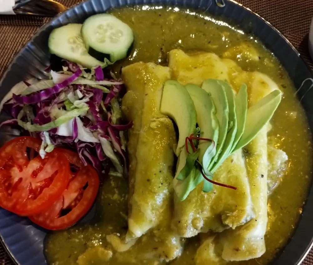 Enchiladas en salsa verde con láminas de aguacate, jitomate y col morada, en un plato oscuro.